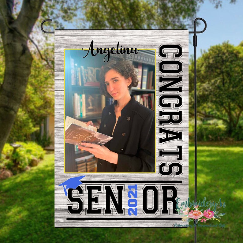 May include: A white wooden sign with a photo of a person holding a book. The sign says "CONGRATS" in large black letters and "SENIOR 2021" in black letters with a blue graduation cap. The sign also says "Angelina" at the top.