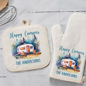 May include: A set of two white cotton pot holders with a watercolor design of a camper in a forest. The pot holders have the text "Happy Campers" and "The Andersons" printed on them.