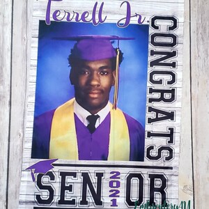 May include: A graduation banner with a photo of a graduate wearing a purple cap and gown. The banner says "Terrell Jr. Congrats Senior 2021" in black and white text with a purple graduation cap graphic.