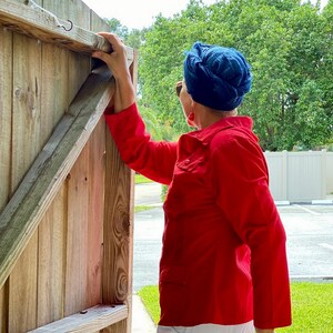 May include: A person wearing a red shirt and a blue turban is opening a wooden gate. The gate is made of vertical wooden planks and has a metal hook at the top.