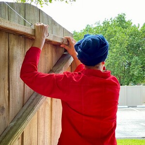 May include: A person wearing a red shirt and a blue headscarf is reaching up to close a wooden gate.