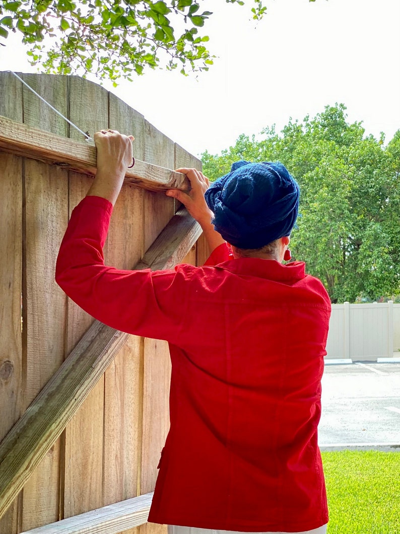 May include: A person wearing a red shirt and a blue headscarf is reaching up to close a wooden gate.