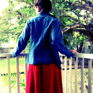 May include: A woman wearing a blue denim jacket and a red skirt stands on a porch with a white railing. She is looking off to the side.