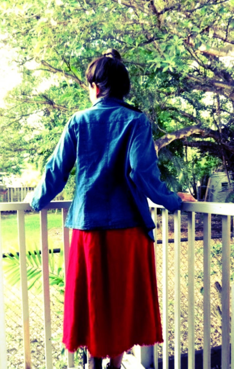 May include: A woman wearing a blue denim jacket and a red skirt stands on a porch with a white railing. She is looking off to the side.