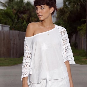 May include: A white top featuring an off-the-shoulder neckline, short sleeves with intricate white crochet lace detailing, and a tie-front hem. The garment includes a subtle embroidered floral pattern on the chest and is paired with white pants.