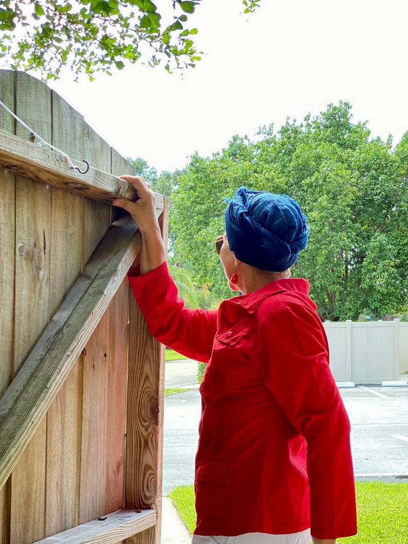 May include: A person wearing a red shirt and a blue turban is opening a wooden gate. The gate is made of vertical wooden planks and has a metal hook at the top.