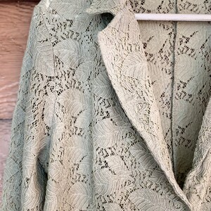 May include: A sage green lace jacket with a long sleeve and a collar. The jacket is made of a delicate lace fabric with a floral pattern.