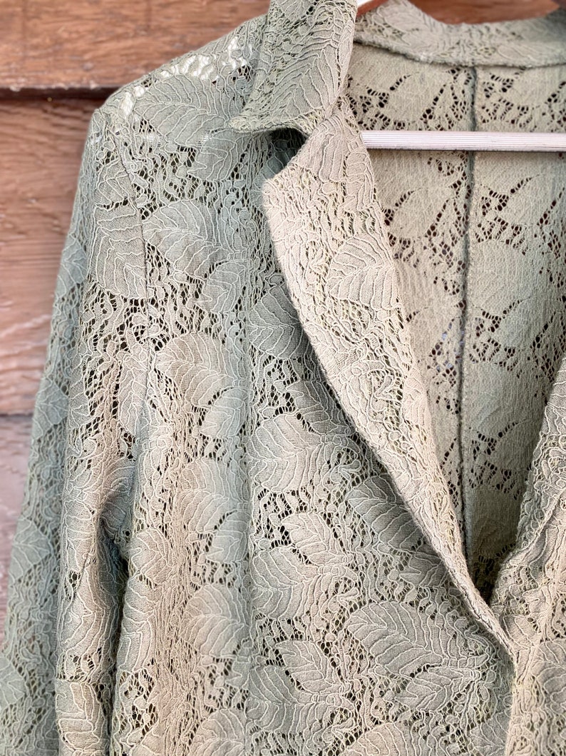 May include: A sage green lace jacket with a long sleeve and a collar. The jacket is made of a delicate lace fabric with a floral pattern.