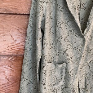May include: A sage green lace blazer with a long sleeve and a pocket on the left side.