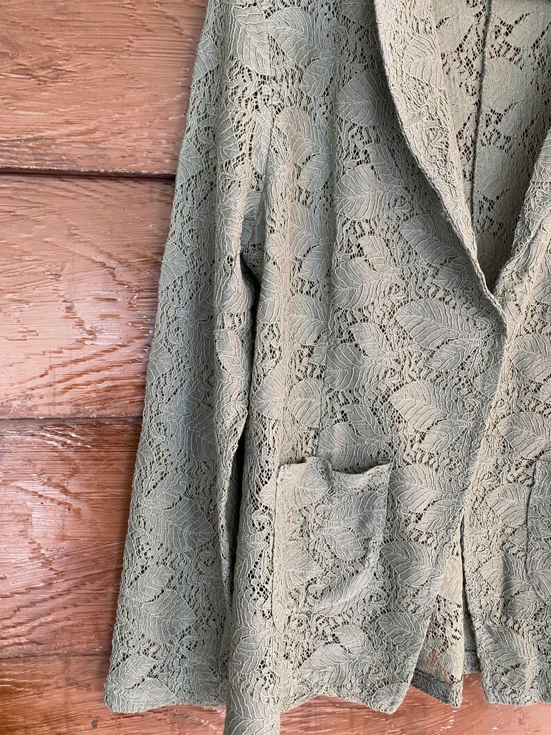 May include: A sage green lace blazer with a long sleeve and a pocket on the left side.