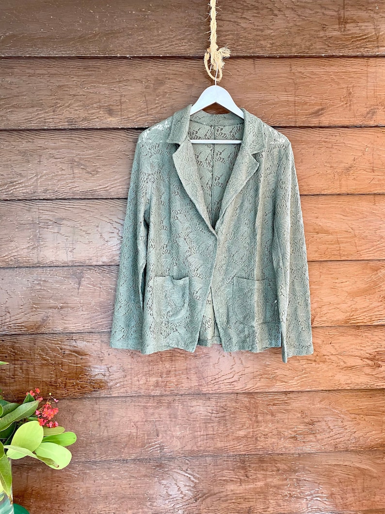 May include: A green crocheted blazer with a button closure and two pockets. The blazer is hanging on a white hanger against a wood background.