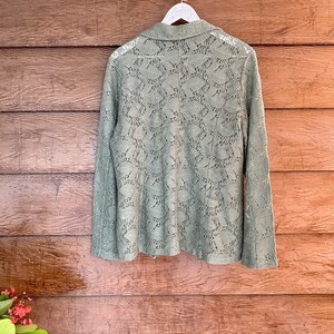 May include: A sage green lace blazer with long sleeves and a collared neckline. The blazer is hanging on a white hanger against a wood background.