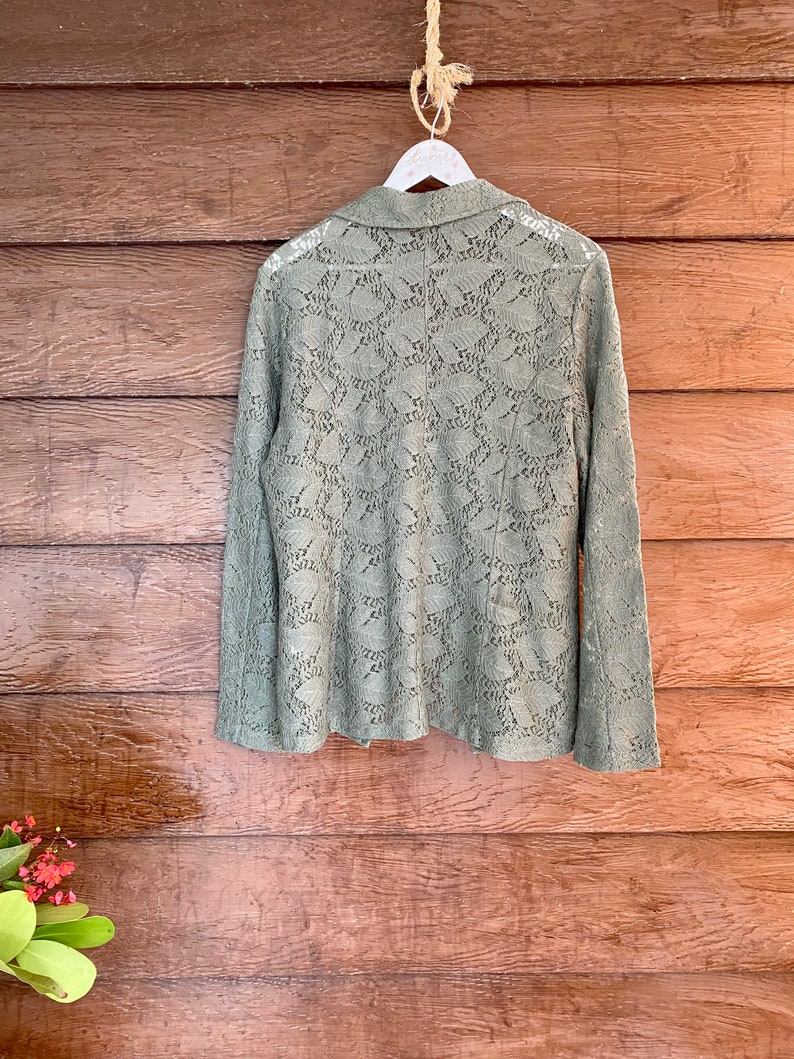 May include: A sage green lace blazer with long sleeves and a collared neckline. The blazer is hanging on a white hanger against a wood background.