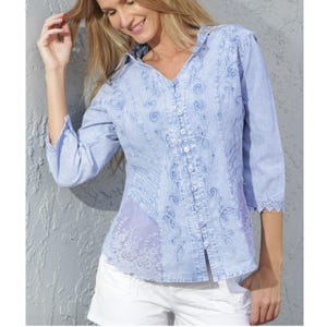 Pode incluir: Camisa azul clara com intrincados bordados florais e em espiral. A camisa apresenta decote em V, mangas três quartos e um painel de renda. É combinada com calções brancos.