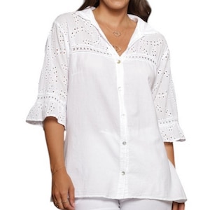Puede incluir: Camisa blanca abotonada con detalles calados en las mangas y el canesú. La camisa tiene cuello camisero y mangas tres cuartos con puños con volantes. La tela parece ligera y transpirable, adecuada para climas cálidos.