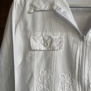 May include: A white jacket with a zipper closure and embroidered floral details. The jacket has a buttoned pocket on the left side.