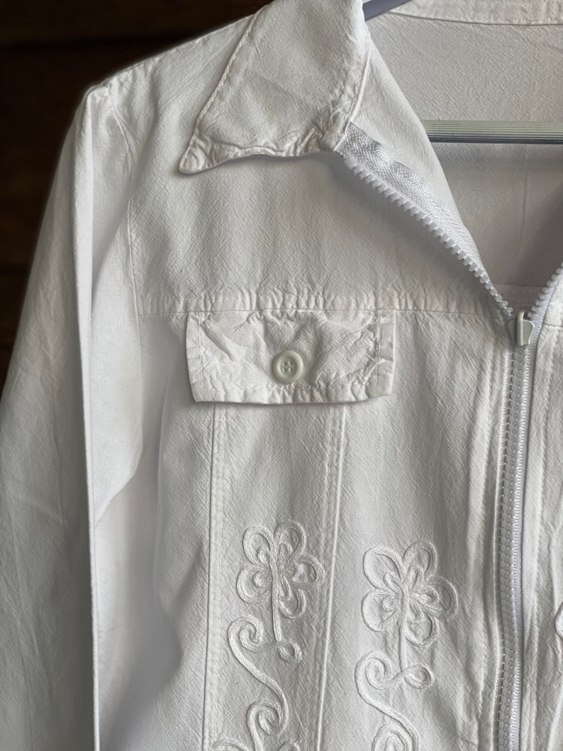 May include: A white jacket with a zipper closure and embroidered floral details. The jacket has a buttoned pocket on the left side.