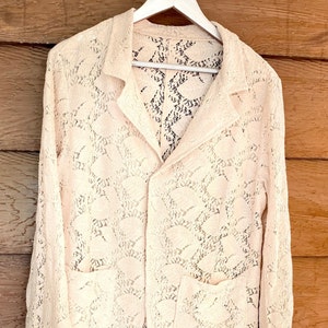 May include: A white lace jacket with a button closure and two pockets. The jacket is hanging on a white hanger against a wooden background.