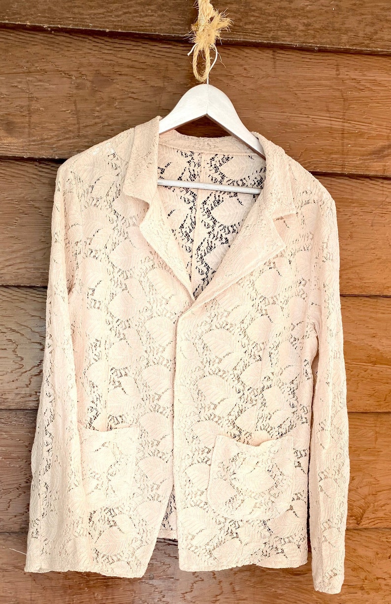 May include: A white lace jacket with a button closure and two pockets. The jacket is hanging on a white hanger against a wooden background.