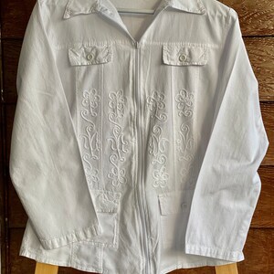 May include: White long-sleeved shirt with a zipper closure and embroidered floral details. The shirt has two flap pockets on the chest and a single pocket on the bottom.