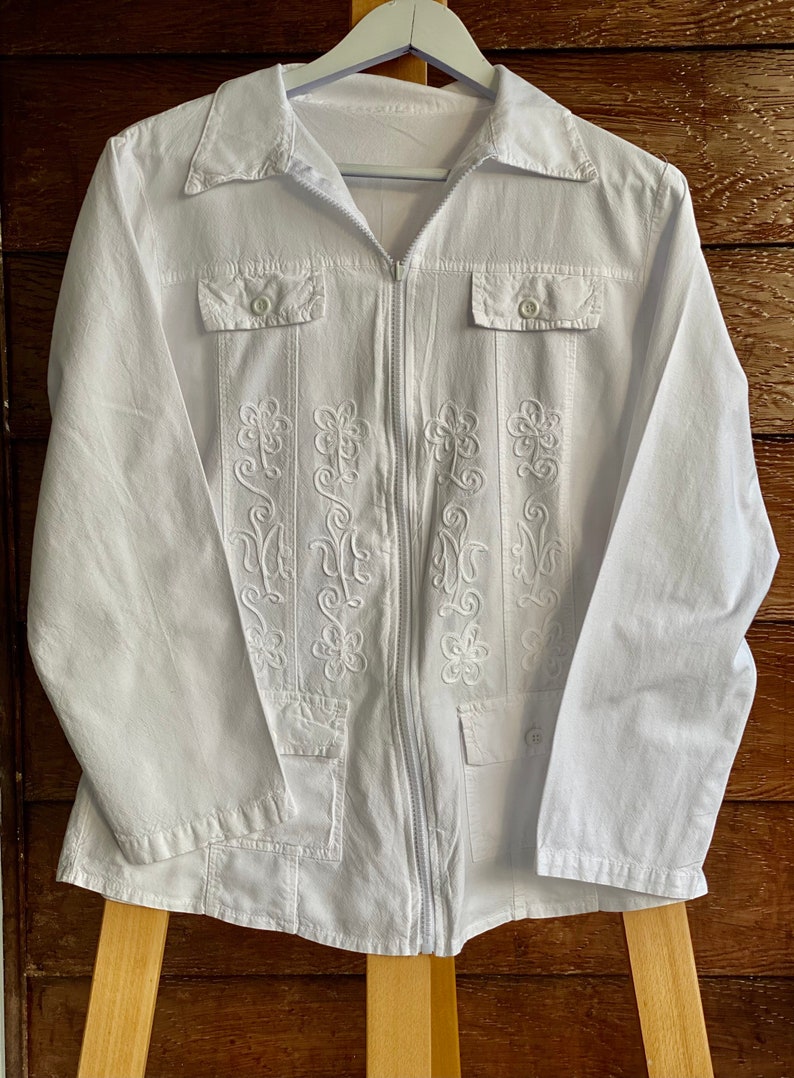 May include: White long-sleeved shirt with a zipper closure and embroidered floral details. The shirt has two flap pockets on the chest and a single pocket on the bottom.