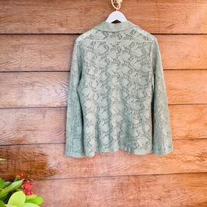 May include: A sage green lace blazer hanging on a white hanger against a wood background.