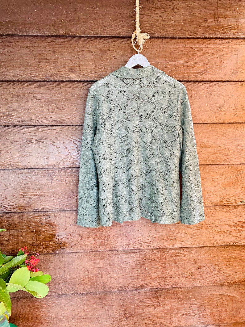 May include: A sage green lace blazer hanging on a white hanger against a wood background.