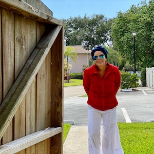 May include: A woman wearing a red shirt, white pants, blue sunglasses, and a blue hat is standing in front of a wooden gate. She is smiling and looking at the camera.