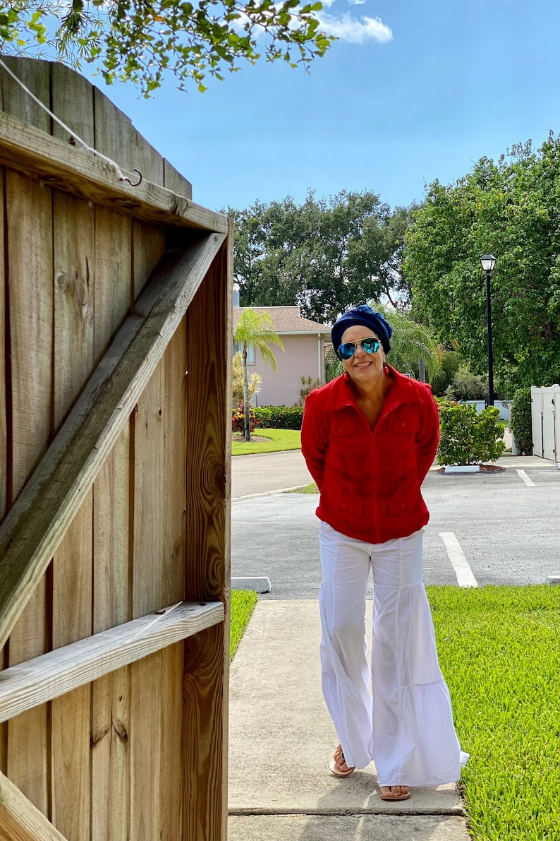 May include: A woman wearing a red shirt, white pants, blue sunglasses, and a blue hat is standing in front of a wooden gate. She is smiling and looking at the camera.