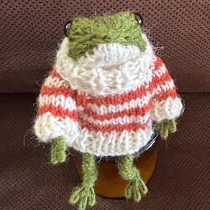 May include: A green knitted frog wearing a white and orange striped sweater. The frog is sitting on a brown surface.