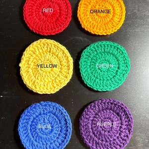 May include: Six crocheted circles in the colours of the rainbow. The circles are red, orange, yellow, green, blue, and purple. Each circle has the colour name written in white in the centre.
