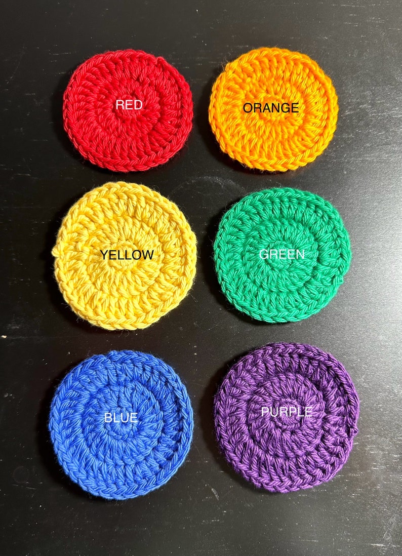 May include: Six crocheted circles in the colours of the rainbow. The circles are red, orange, yellow, green, blue, and purple. Each circle has the colour name written in white in the centre.