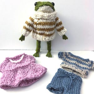 May include: A green knitted frog doll wearing a white and tan striped sweater, accompanied by a separate lavender knitted dress with a white beaded neckline, a blue and white striped shirt, and a pair of blue knitted shorts with a drawstring.