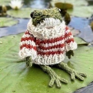 May include: A small, green, crocheted frog figurine wearing a thick, hand-knitted sweater with horizontal red and white stripes. The frog has dark, round eyes and textured, yarn-based skin, sitting upright with its long, spindly legs extended.
