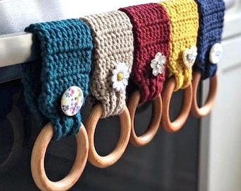 May include: Five crochet towel holders in teal, beige, burgundy, mustard yellow, and navy blue. Each features a wooden ring at the bottom and a decorative button in the center, including floral, daisy, and crystal-like designs, hanging on a handle.