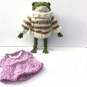 May include: A green knitted frog doll with black button eyes, wearing a cream and brown striped sweater. Beside the frog is a separate lavender knitted dress featuring a white beaded neckline, designed for the doll to wear as an accessory.
