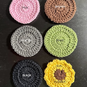 May include: Six crocheted coasters in various colours and designs. The coasters are round, except for one which is a sunflower shape. The colours are pink, brown, grey, green, black, and yellow. The coasters have the words "PINK", "BROWN", "PEWTER", "SAGE", "BLACK", "SUN", and "FLOWER" written on them.