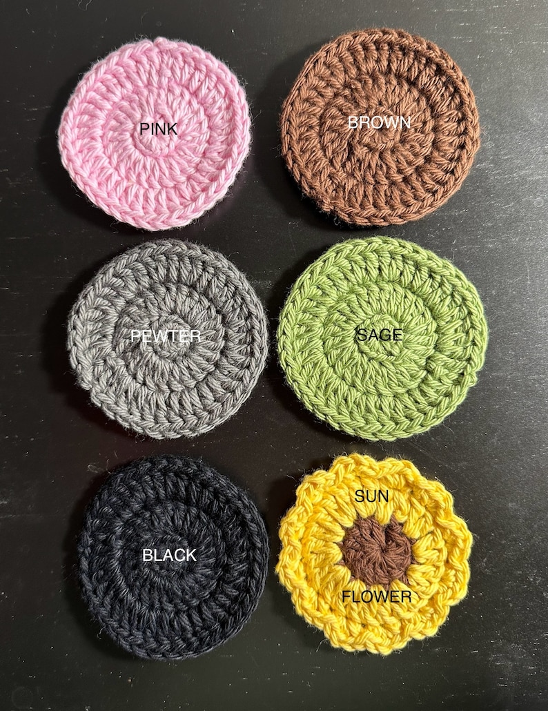 May include: Six crocheted coasters in various colours and designs. The coasters are round, except for one which is a sunflower shape. The colours are pink, brown, grey, green, black, and yellow. The coasters have the words "PINK", "BROWN", "PEWTER", "SAGE", "BLACK", "SUN", and "FLOWER" written on them.