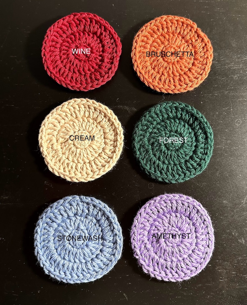 May include: Six crocheted coasters in different colours and patterns. The colours are wine, bruschetta, cream, forest, stonewash, and amethyst. The coasters are all round and have a raised centre.