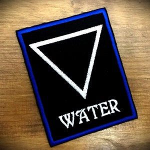 Może przedstawiać: Czarna naszywka haftowana z białym trójkątem i napisem "WATER" białymi literami. Naszywka ma niebieski obramowanie.