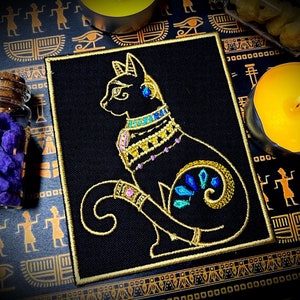 Op de afbeelding: Gestikt patch met een zwarte kat met gouden en blauwe versieringen. De kat is afgebeeld in een gestileerde Egyptische stijl, met een kroon en sieraden. Het patch is op een zwarte achtergrond.