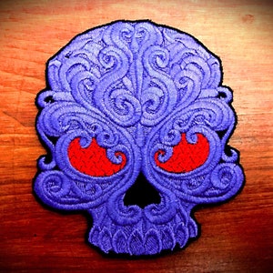 Peut inclure: Écusson brodé en forme de crâne avec des volutes violettes complexes et des orbites rouges. Le crâne a un nez noir et est posé sur un fond en bois.