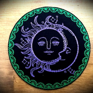 Op de afbeelding: Een geborduurde zon- en maanontwerp in paars en wit op een zwarte stoffen patch met een groene geborduurde rand.