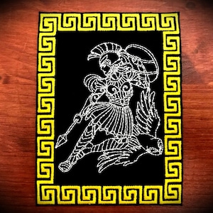 May include: A black embroidered patch featuring a white outline of a Greek warrior holding a spear and shield. The patch is bordered by a yellow Greek key design.