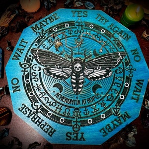 Könnte beinhalten: Ein blaues Ouija-Brett aus Holz mit einem schwarz-weißen Motten-Design. Das Brett hat die Wörter "JA", "NEIN", "VIELLEICHT", "NOCHMAL VERSUCHEN", "WARTEN" und "UMFORMULIEREN" am Rand gedruckt. Das Brett hat auch einen Kreis in der Mitte mit verschiedenen Symbolen und den Worten "ACHERONTIA ATROPOS".