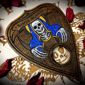 May include: Wooden planchette with a dark brown background and a detailed illustration of a skeleton wearing a blue and yellow robe. The text "SANTA MUERTE" is written in a decorative font above the skeleton.