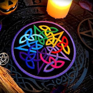 Peut inclure: Un patch en tissu noir avec une bordure violette, brodé d'un motif de nœud celtique aux couleurs de l'arc-en-ciel. Le patch est entouré d'une surface en bois foncé avec un pentagramme et d'autres éléments décoratifs.