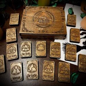 Peut inclure: Une boîte en bois contenant un jeu de cartes de tarot. Les cartes présentent divers symboles et illustrations, notamment une vague, un oiseau, une étoile, un soleil, une récolte, une femme, une faucille, un anneau et un carrefour. La boîte est décorée de sculptures complexes et a un couvercle avec un motif de triangle avec une flamme à l'intérieur.