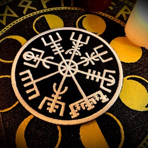 Può includere: Una toppa ricamata in bianco e nero con un design a bussola. La bussola ha quattro punti cardinali contrassegnati da simboli. La toppa è su uno sfondo nero con un motivo dorato.