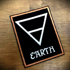 Könnte beinhalten: Schwarz-weiß gestickter Patch mit einem weißen Dreieckssymbol und dem Wort "EARTH" in weißen Buchstaben. Der Patch hat einen braunen Rand.